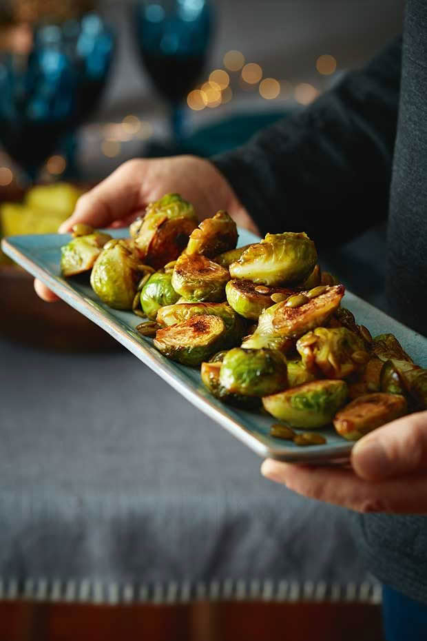 A person carries a long plate with honey roasted brussels sprouts and pepitas. 