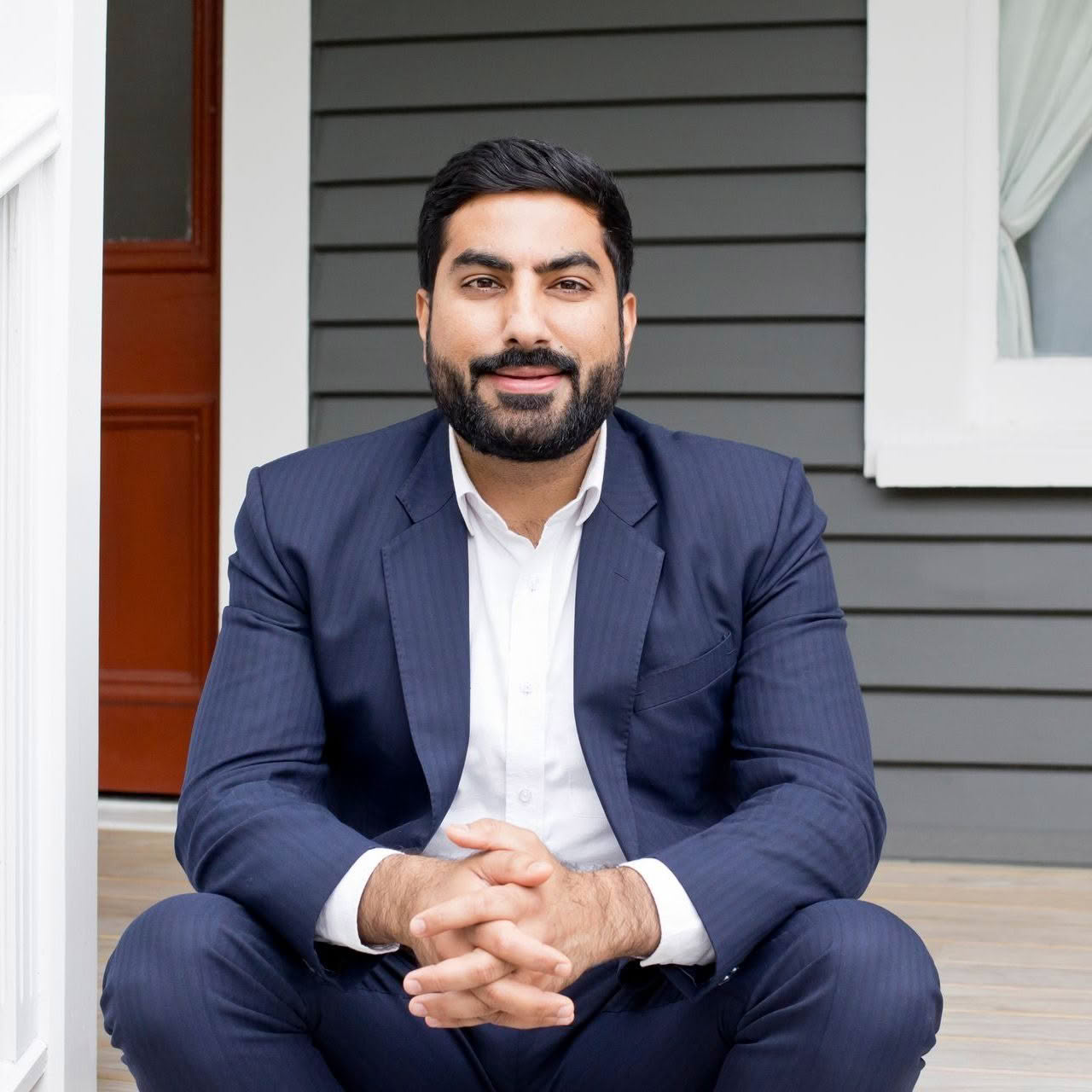 A man in a suit is sitting on a porch with his hands folded