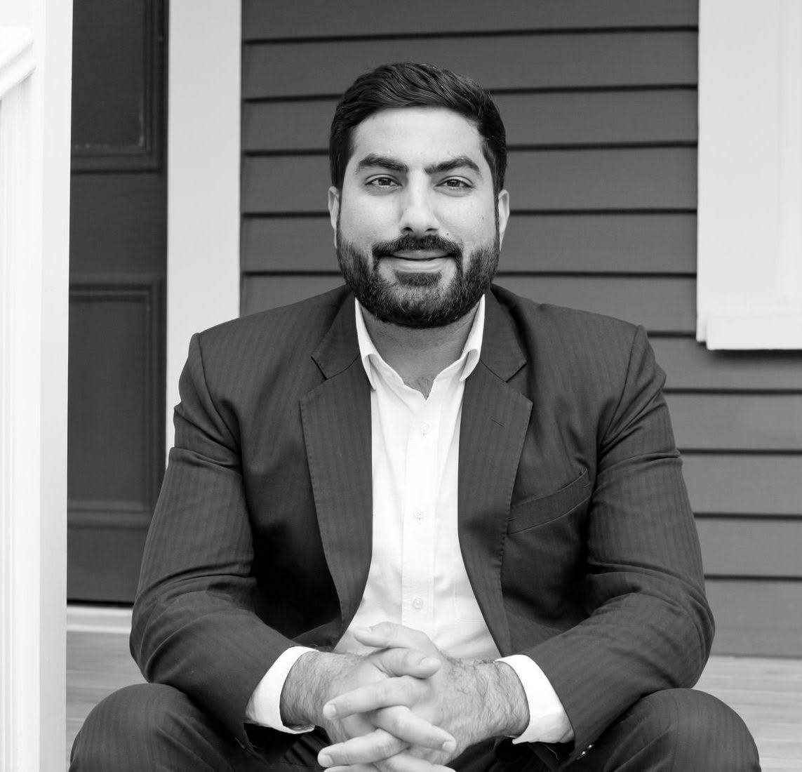 A man in a suit is sitting on a porch with his hands folded.