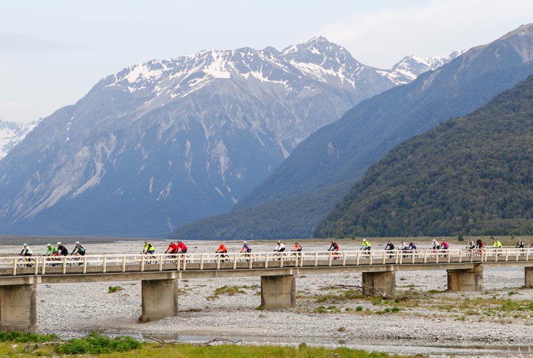 A group of people are riding bikes on a bridge over a river with mountains in the background.