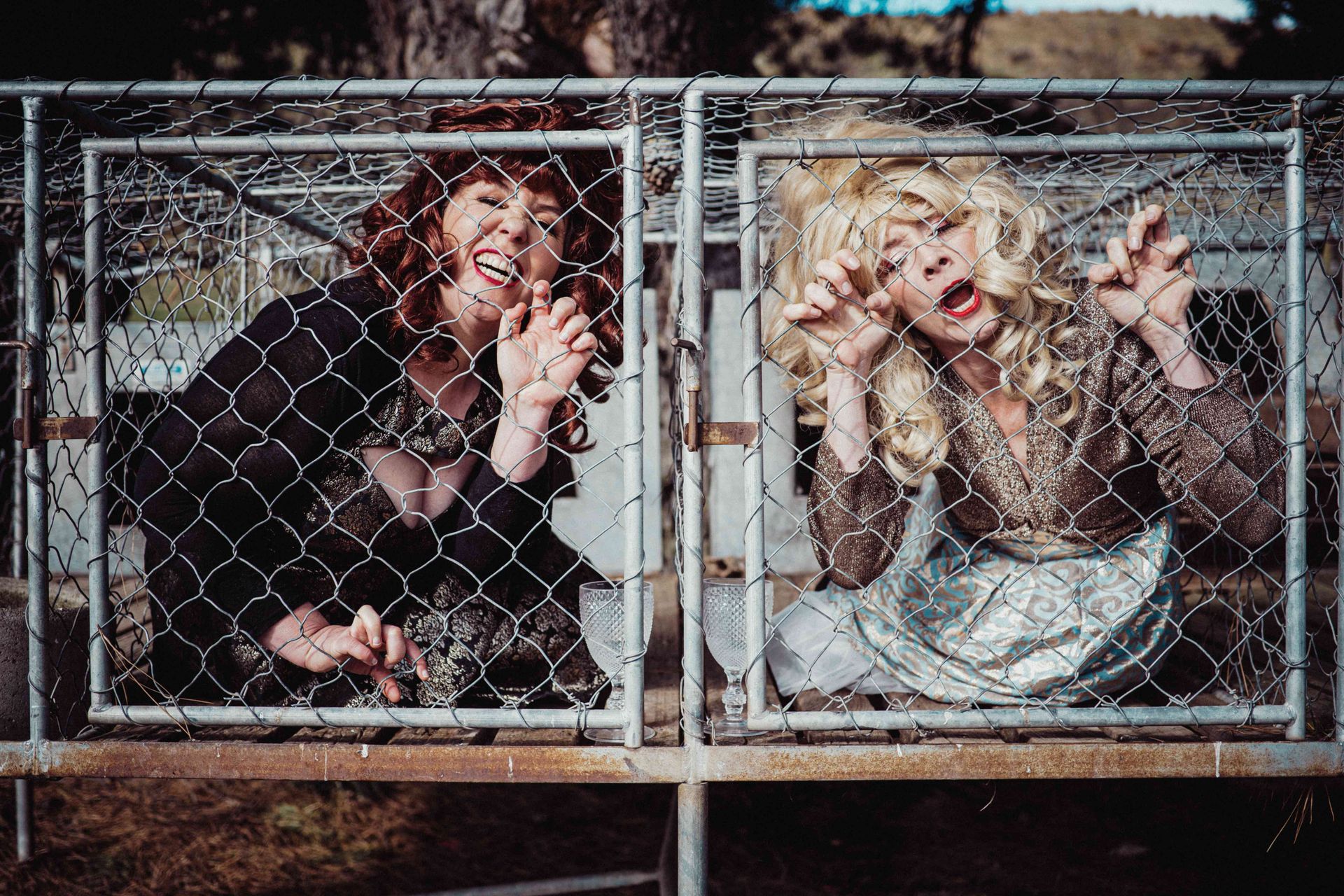 Two women in wigs are behind a chain link fence.