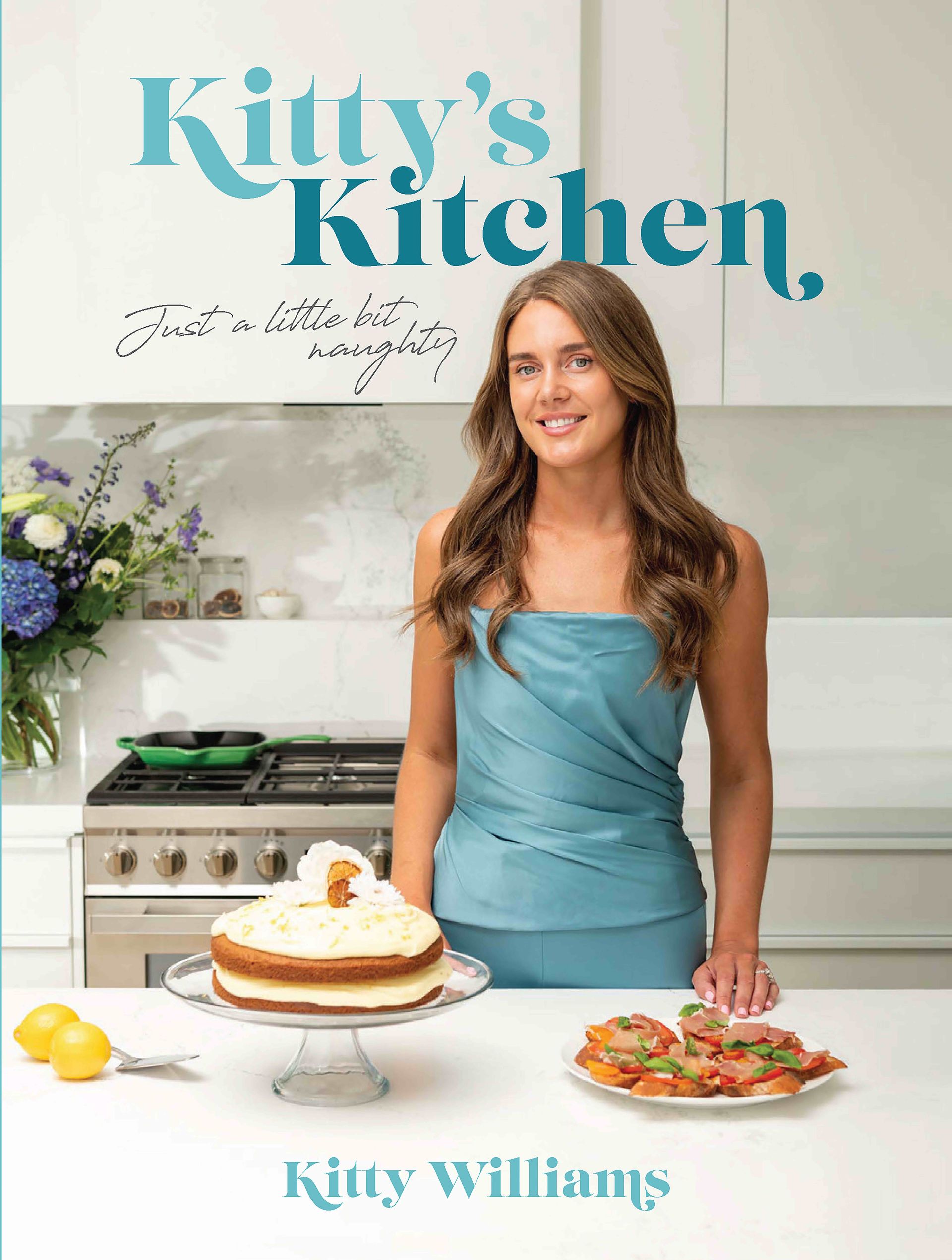 A woman in a blue dress is standing in a kitchen next to a cake.