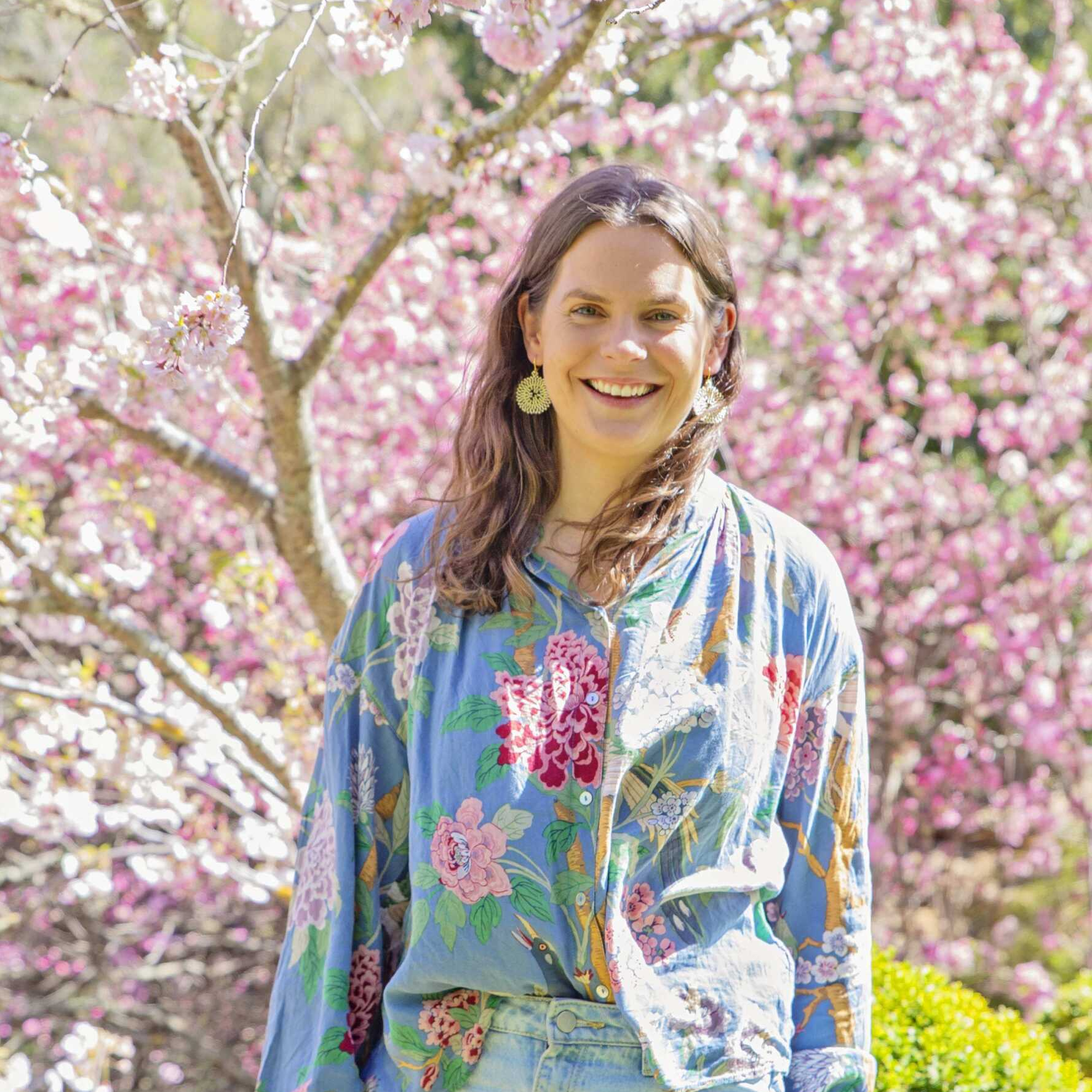 A woman in a blue floral shirt is standing in front of a tree with pink flowers.
